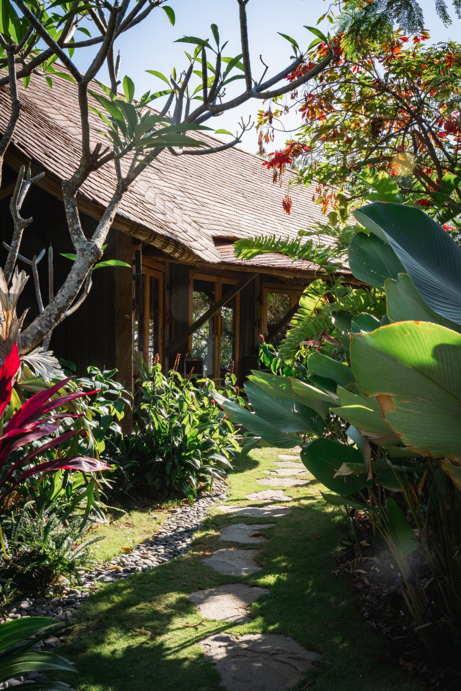 Stone path leading through lush tropical gardens to a wooden yoga shala under sunlight at a Bali retreat.