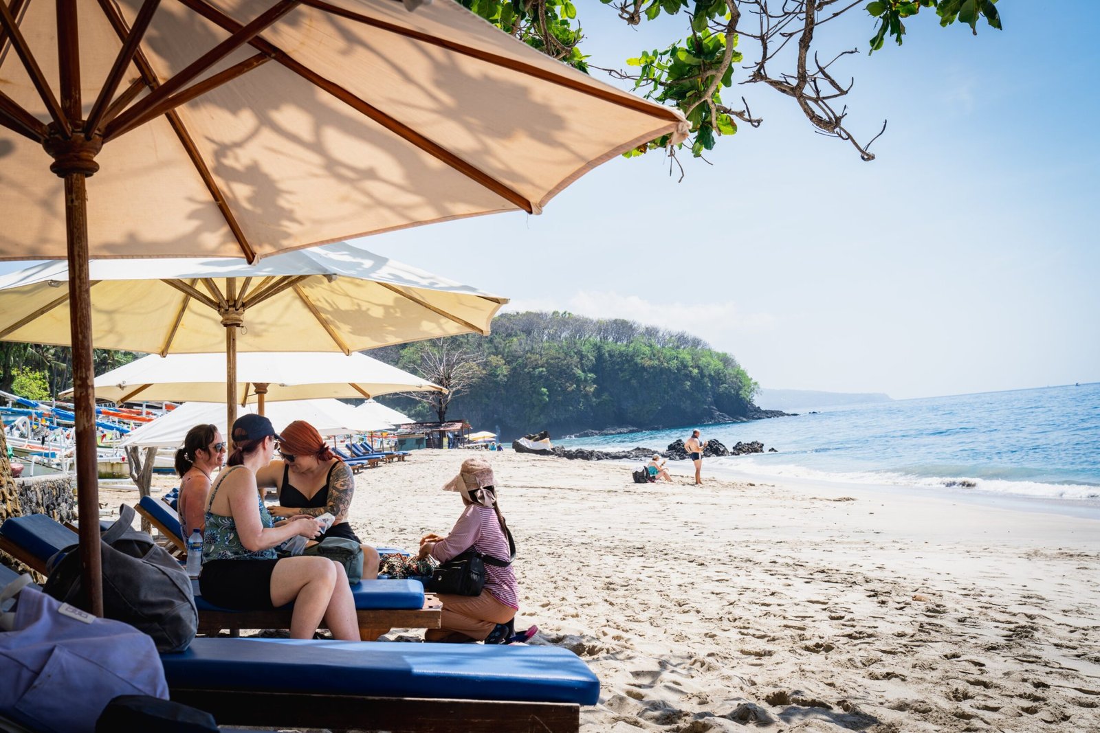 Guests relaxing under umbrellas on a white sandy beach with turquoise water at a wellness retreat in Bali.